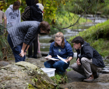 Three people examine papers and a plastic container while sitting on rocks beside a stream; others stand in the background near trees and water.